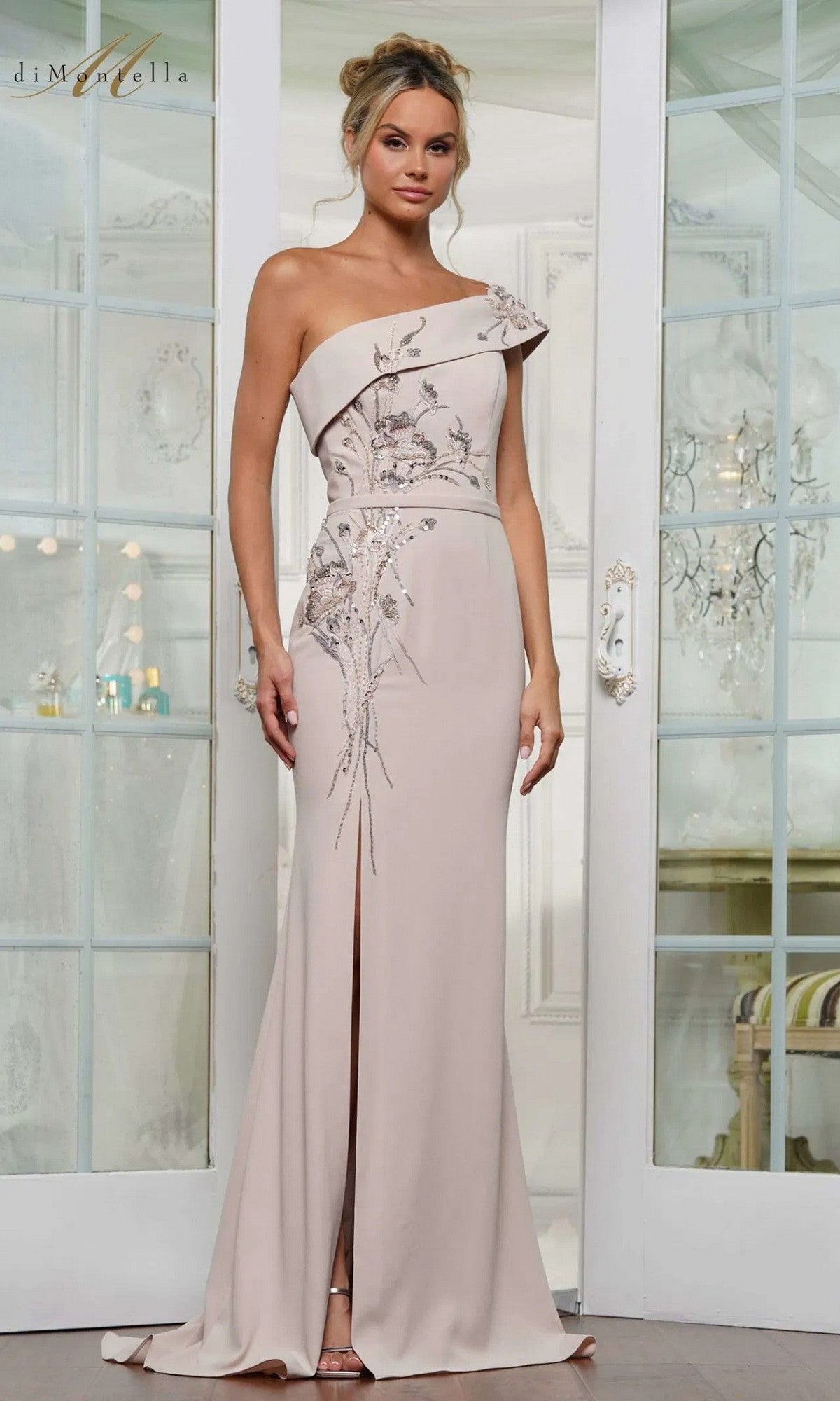 A woman wearing the Rina Di Montella RD3203 long formal dress in champagne, featuring floral embroidery, a one-shoulder design, and a front slit, stands before white double doors.
